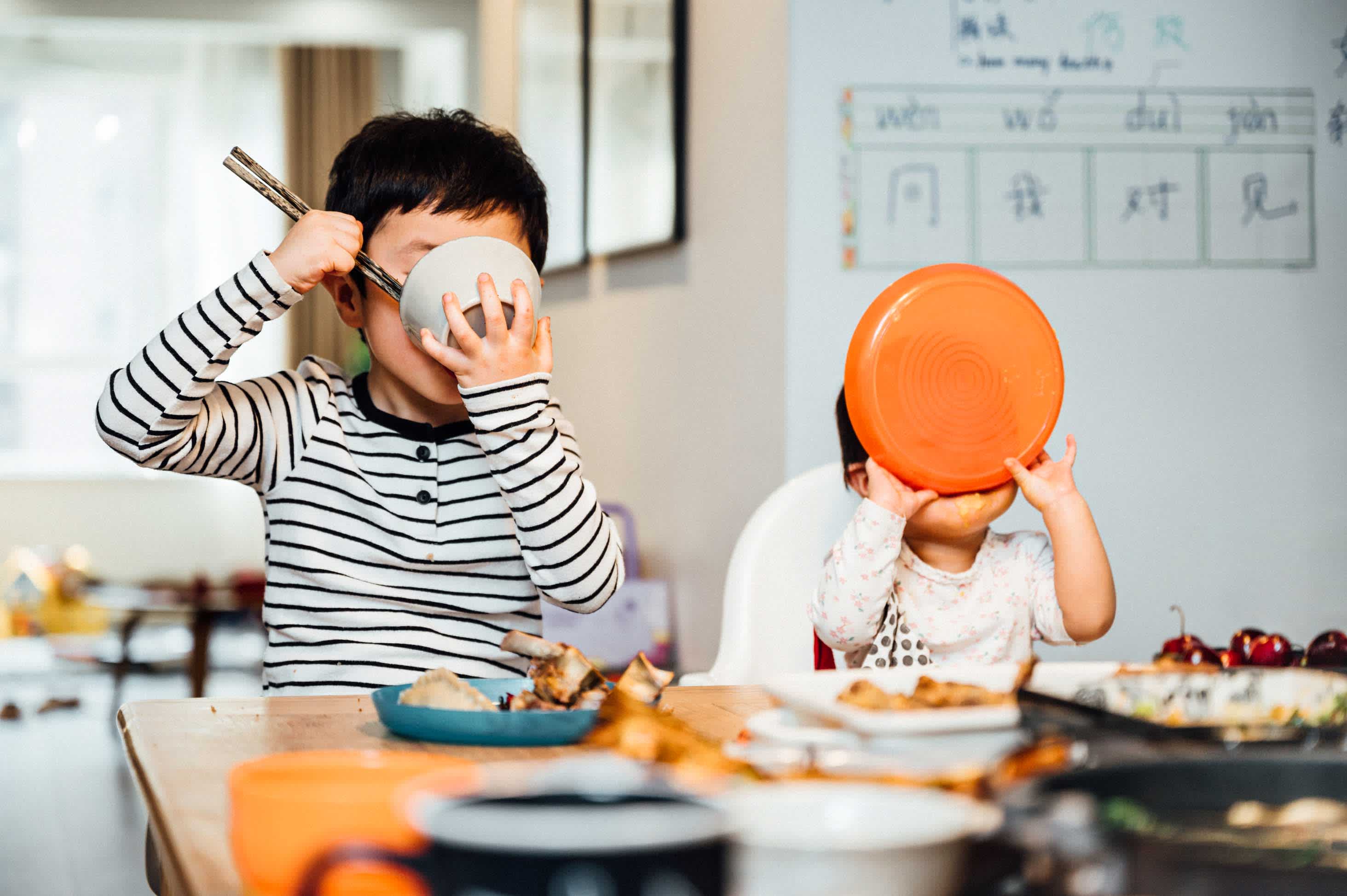 Two young boys sitting side by side at the kitchen table with a decadent spread laid out in front of them. Both have their bowls up to their mouths and are eating every last bite of food.