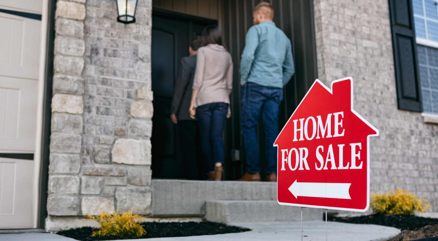 A group of people are about to walk into a home that has a for sale sign on the front yard.