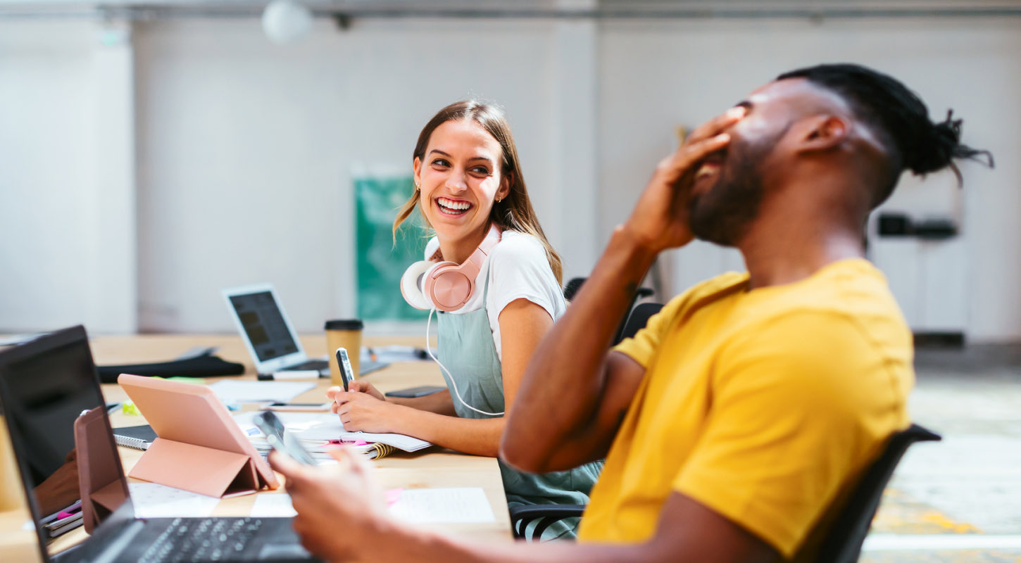 Two young laughing adults are working from their laptops while sitting at a communal desk.