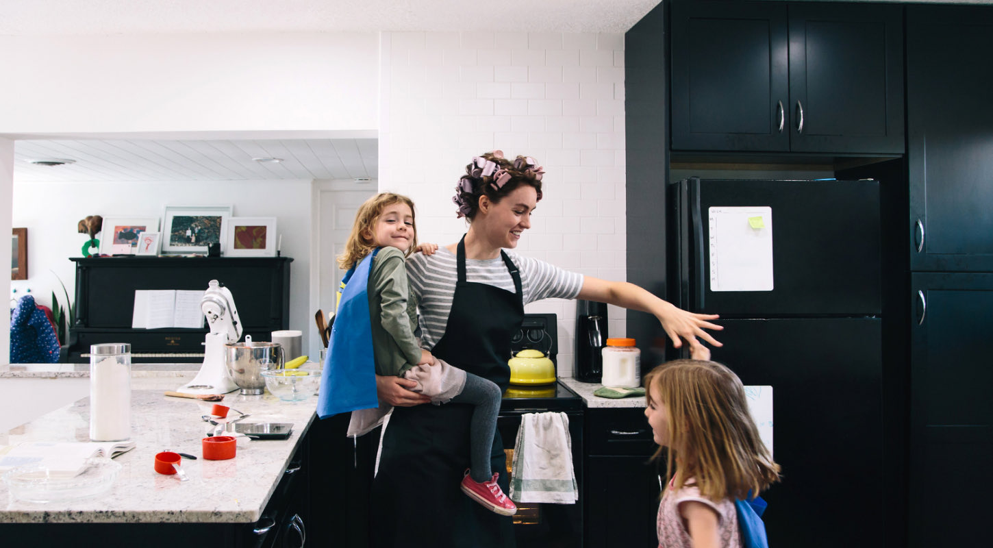 A woman in hair rollers is standing in front of the kitchen counter with her two school-age children. She holds one daughter in her arms and twirls the other by her finger. The counter is scattered with mixing bowls, measuring cups, and flour as if getting ready to bake. 