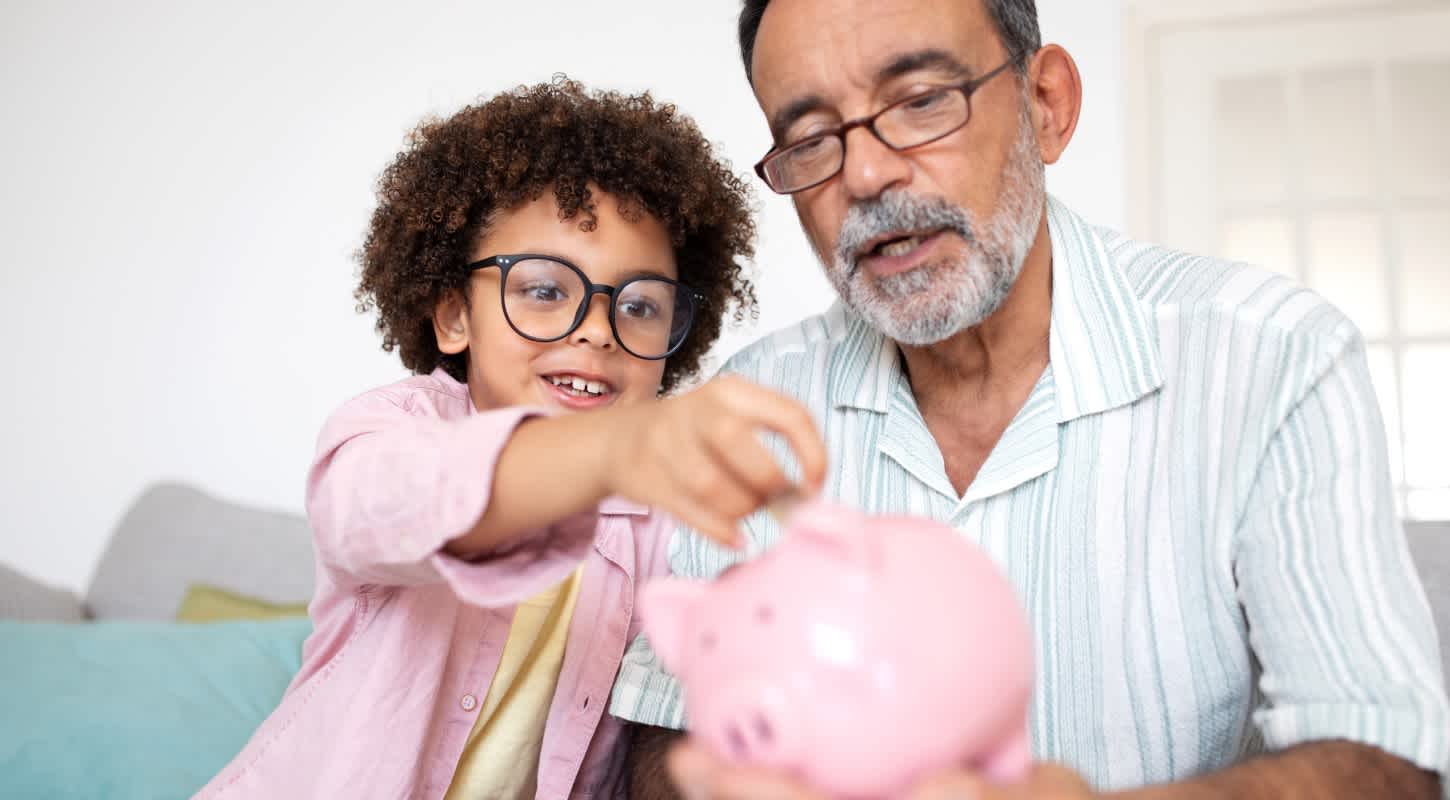 A young boy and his grandfather sit side by side, smiling as they deposit savings into a piggy bank together.