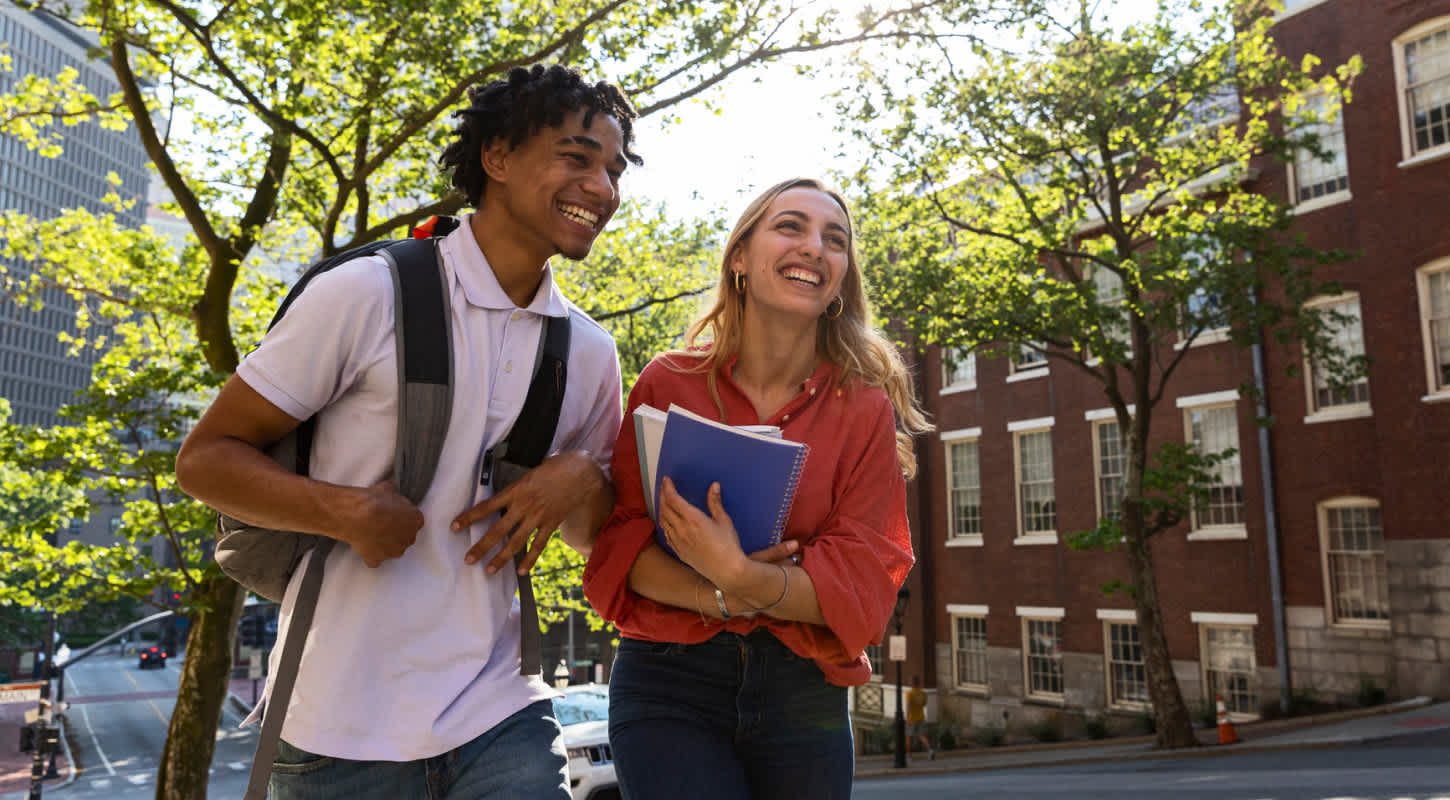 Two young students walk outdoors through an urban college campus with books in hand on a sunny day.
