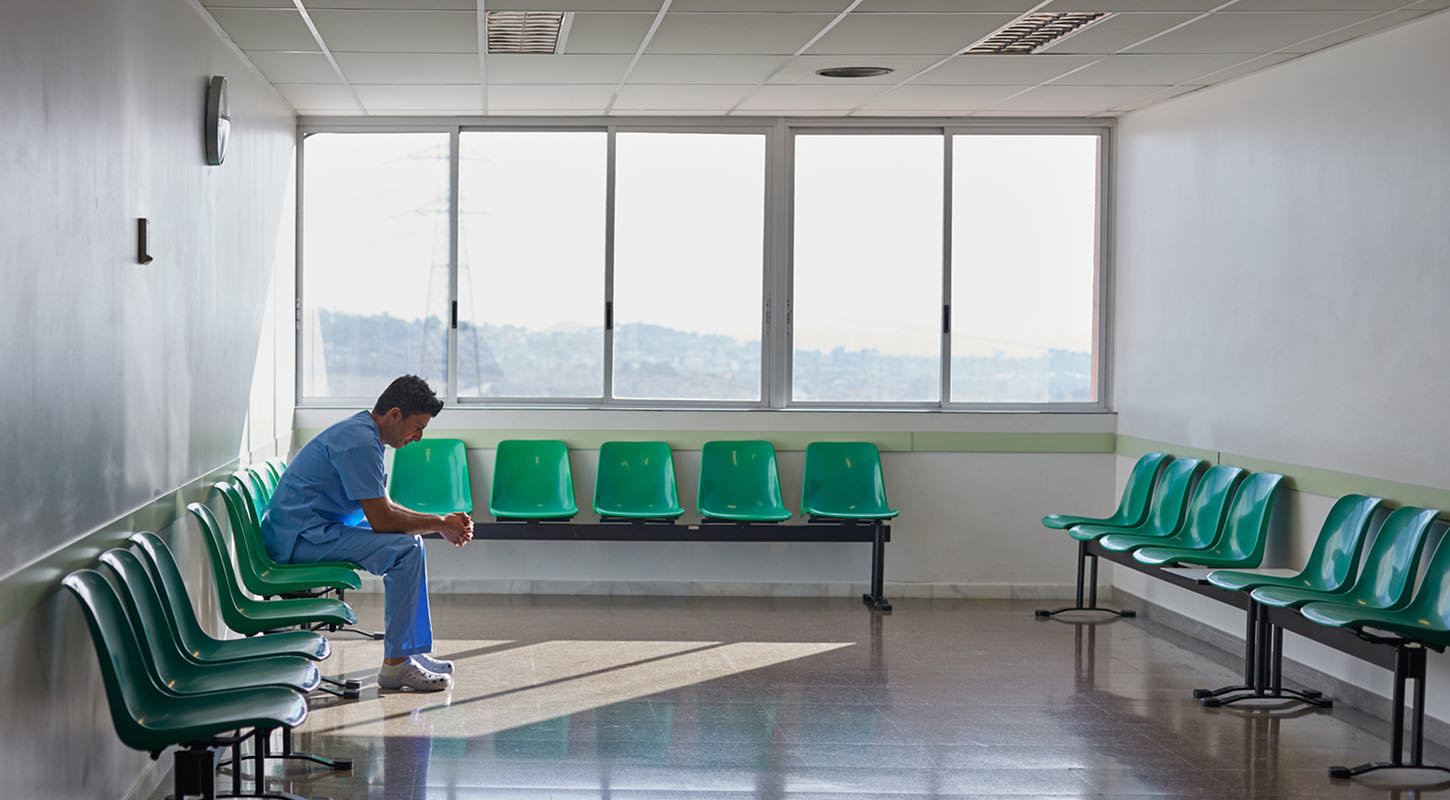 A young man in blue medical scrubs sits pensively in a quiet waiting room, hands clasped together with elbows resting on his knees, eyes gazing downward in thought.