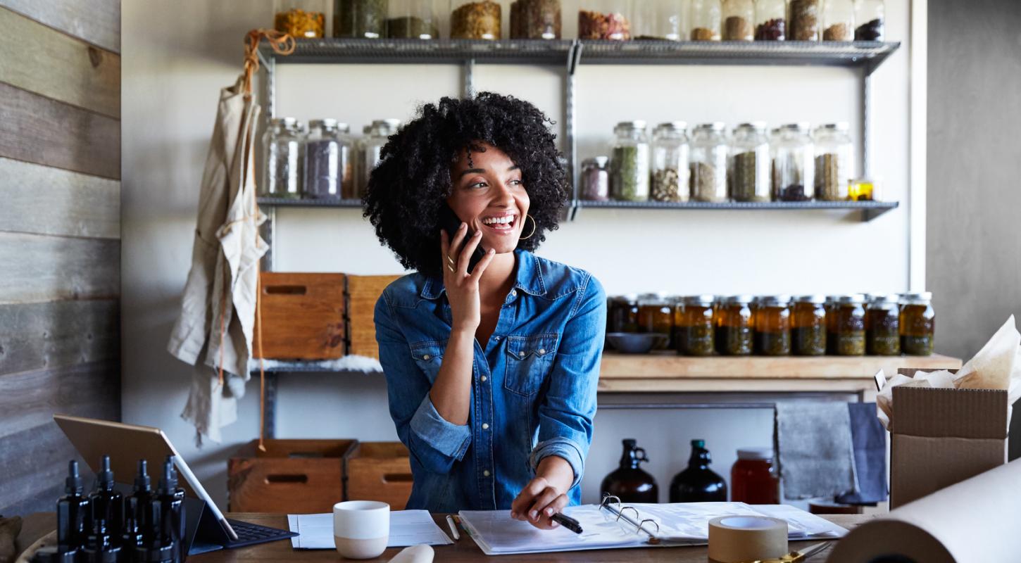 A smiling woman with curly hair, wearing a denim shirt, talks on her phone while working at a wooden counter in a small shop or studio. She gestures with a pen over an open binder, surrounded by packaging materials, a tablet, and dark glass bottles. Behind her, shelves are neatly lined with glass jars filled with herbs, spices, or dried goods, suggesting a natural or handmade product business. The setting is warm, rustic, and organized, reflecting a cozy and entrepreneurial atmosphere.