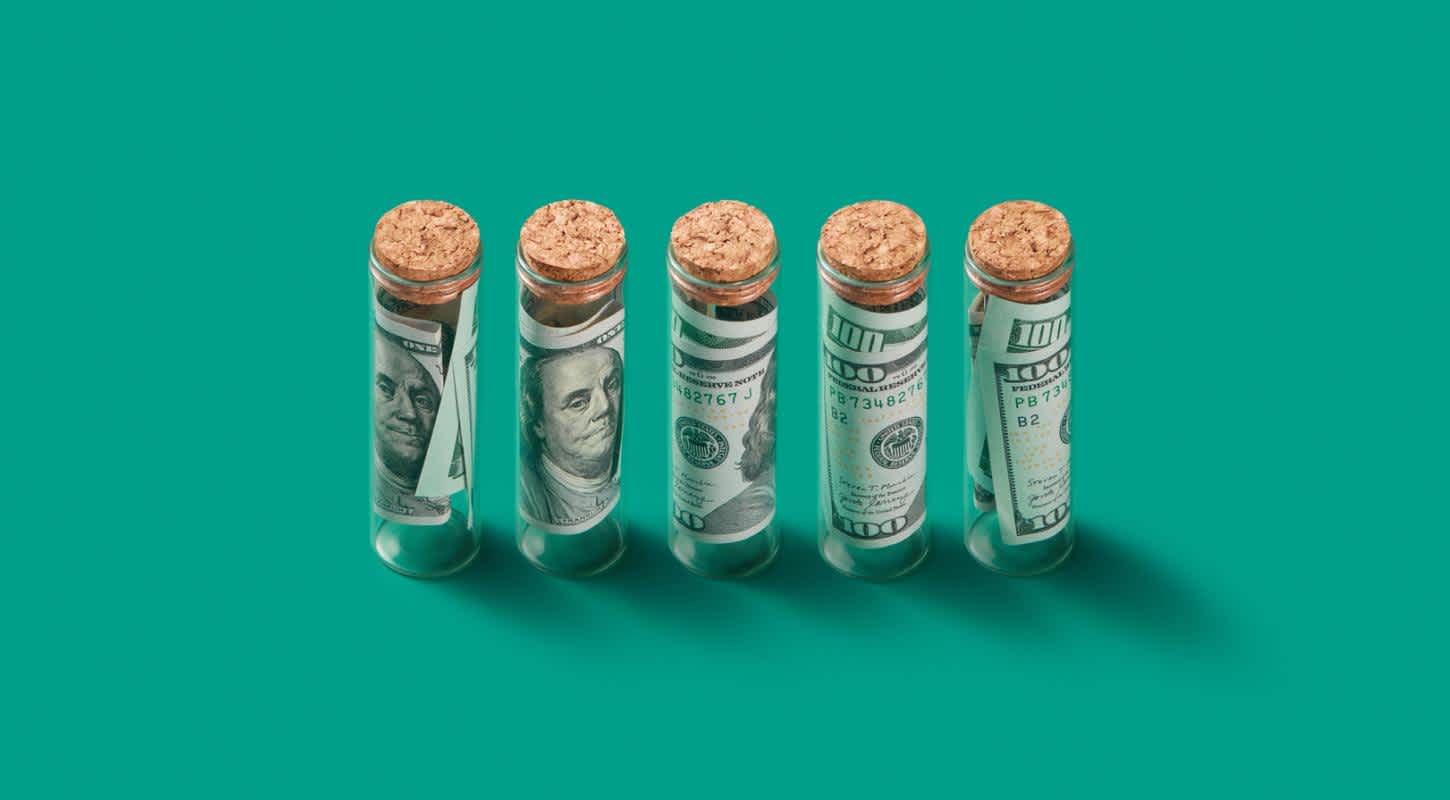 Five small glass vials, each sealed with a cork lid, contain rolled-up $100 bills.