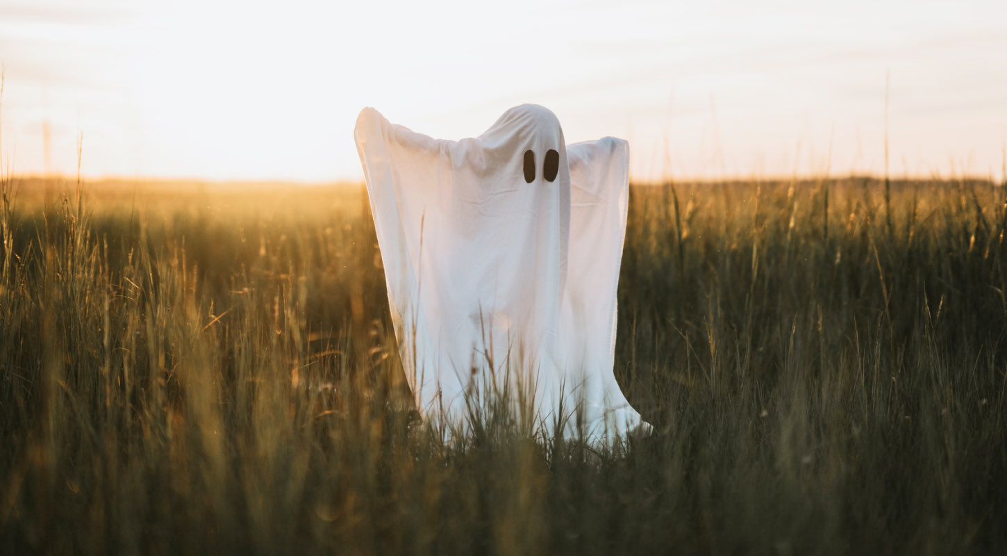 A trick-or-treater stands in the middle of an open field dressed as a spooky ghost. The costume consists of a white sheet with two black oval-shaped eyes. 