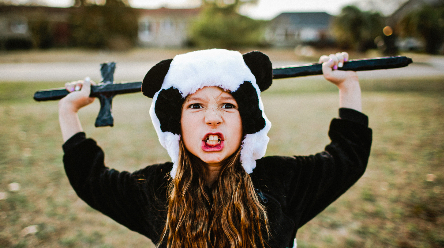 A young girl wearing a panda hat holds a play sword in an overhead press position while gritting her teeth and giving a battle stare.