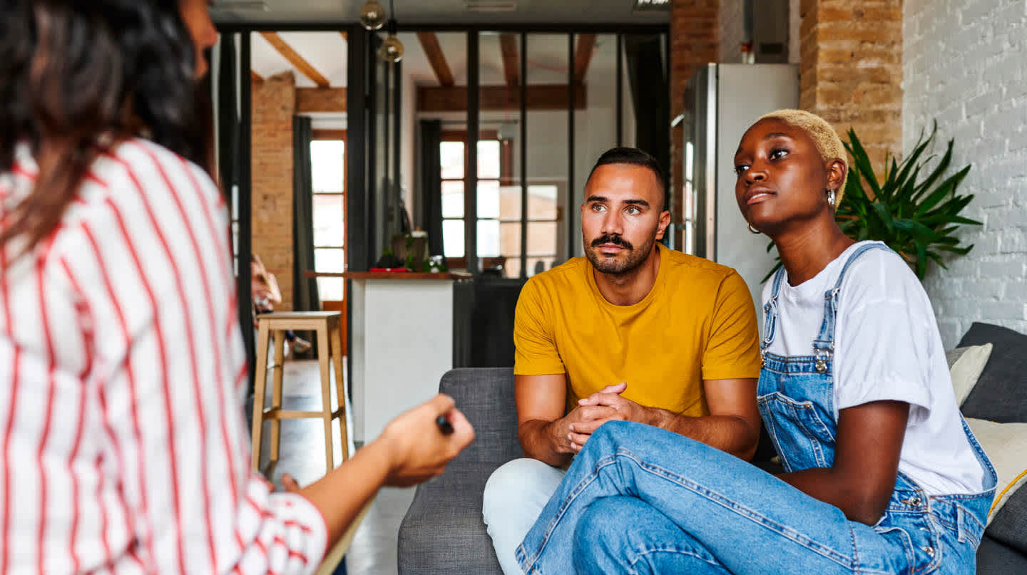 A young couple sitting side by side on a sofa listens intently to guidance being provided by an advisor sitting directly across from them inside of a Brooklyn-style apartment.