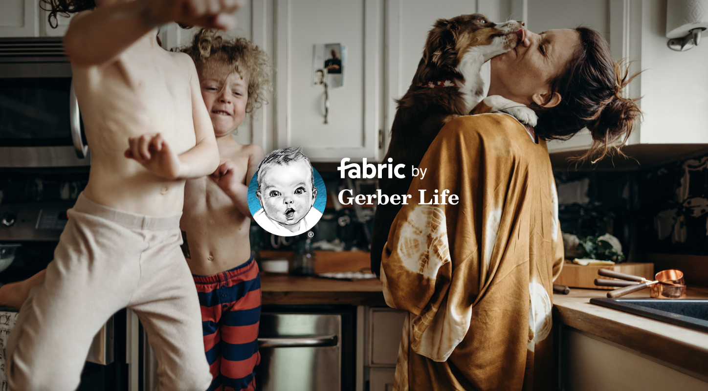 Two toddlers jump around in a kitchen, while their mother is cuddling a dog that is licking her face as she takes a break from cooking a meal. 