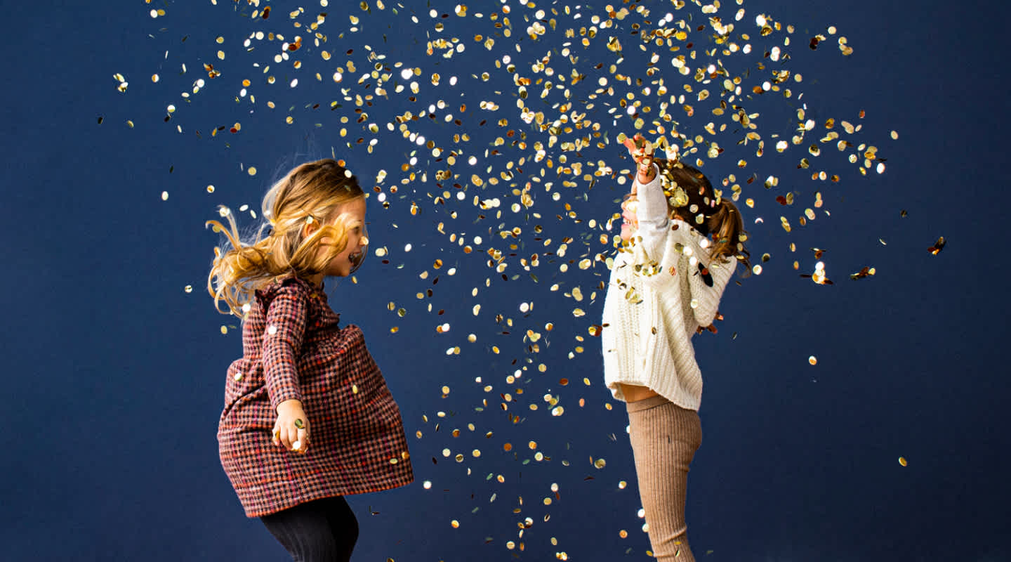 Two young girls dressed in festive attire jump while throwing their hands in the air amidst flying gold confetti.