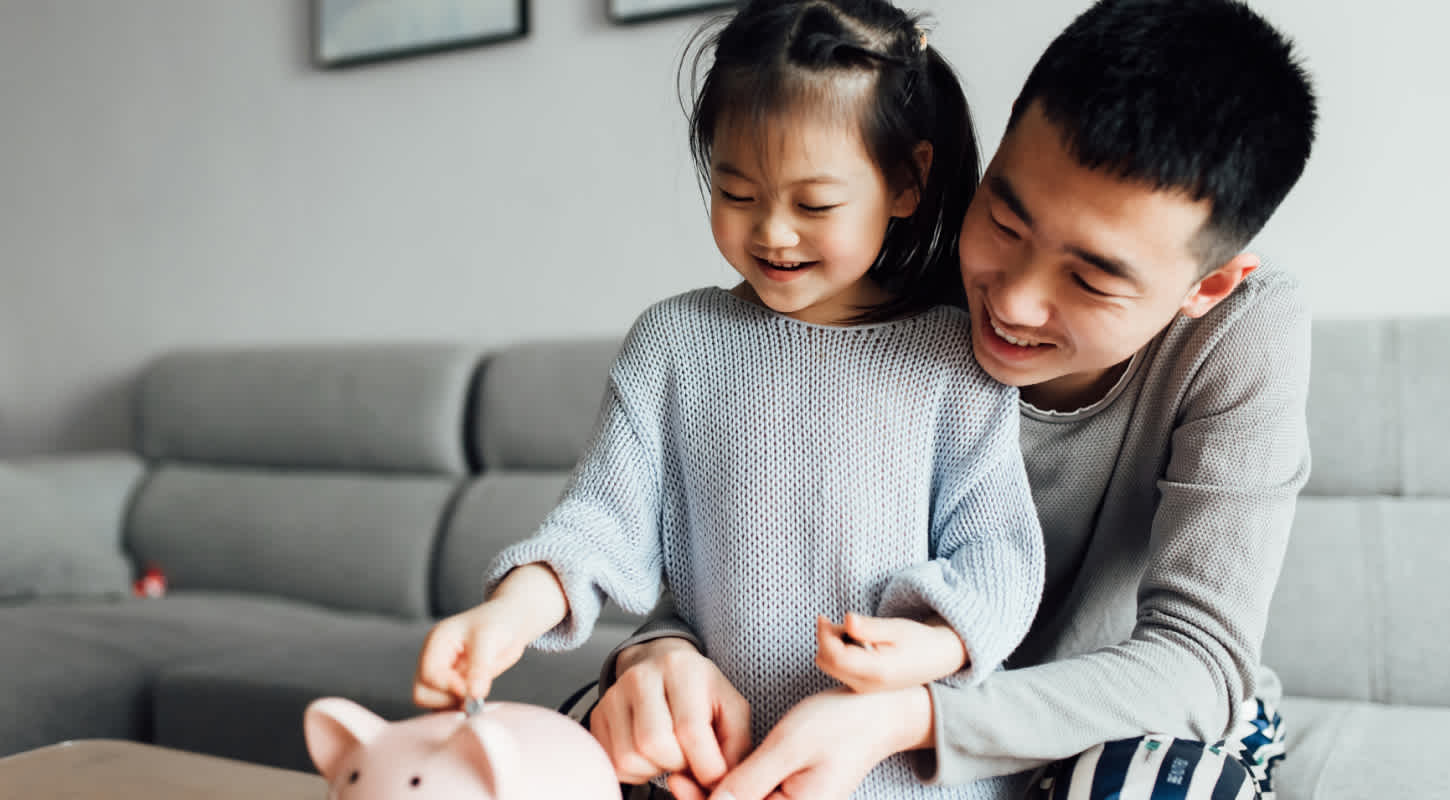 A father embraces his toddler daughter as he observes her place money inside of a piggy bank that sits on a coffee table.
