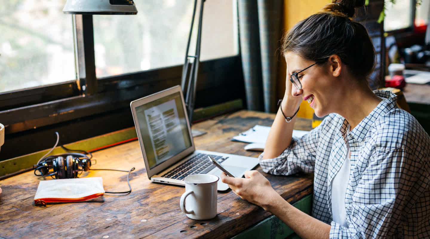 A young woman wearing glasses sits in front of a window at a co-working cafe and gazes at her cell phone in hand, while an open laptop, coffee, headphones, and journal sit ideally in front of her.