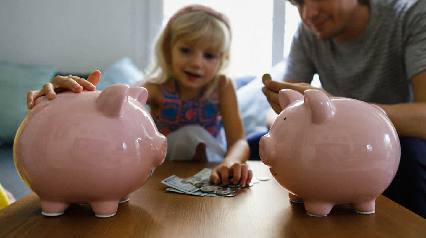 A young girl and her father sit at a coffee table in front of two piggy banks, several dollar bills, and loose change. She places her hand on one of the two piggy banks.