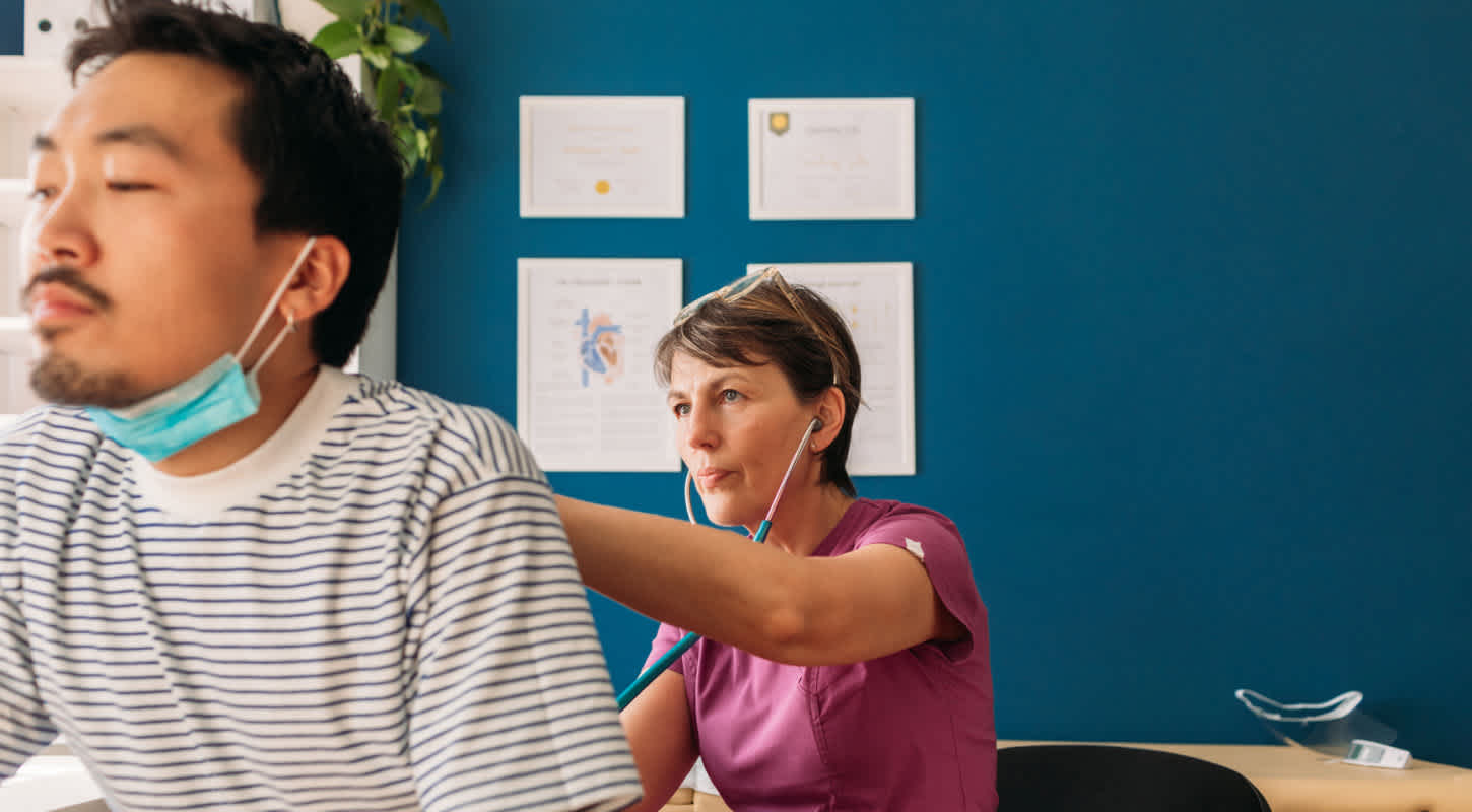 A young patient has his lungs examined by a doctor with a stethoscope. They both wear face masks while she places the diaphragm on his back and listens intently.
