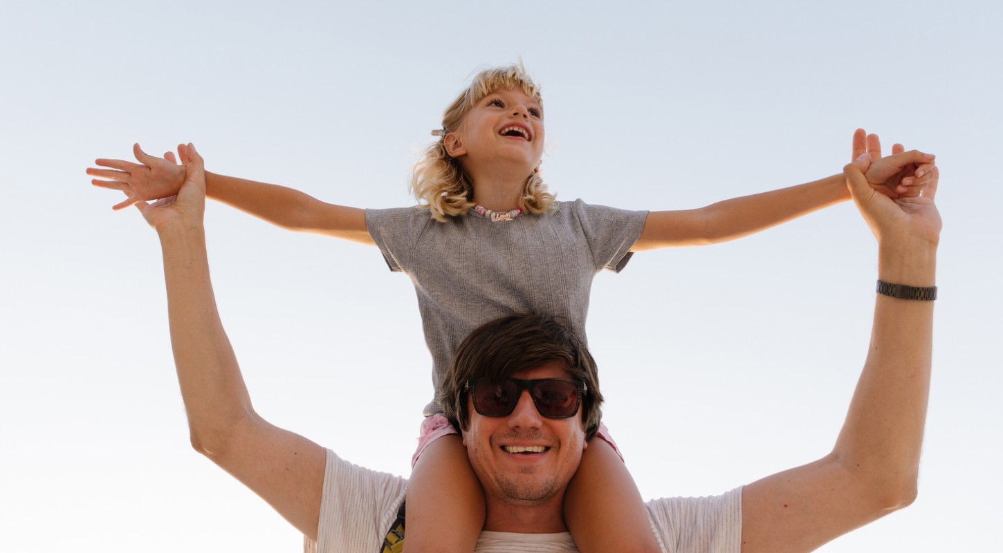 A toddler-aged girl sits atop her father's shoulders, her arms outstretched as he holds her hands. Both are laughing and smiling.