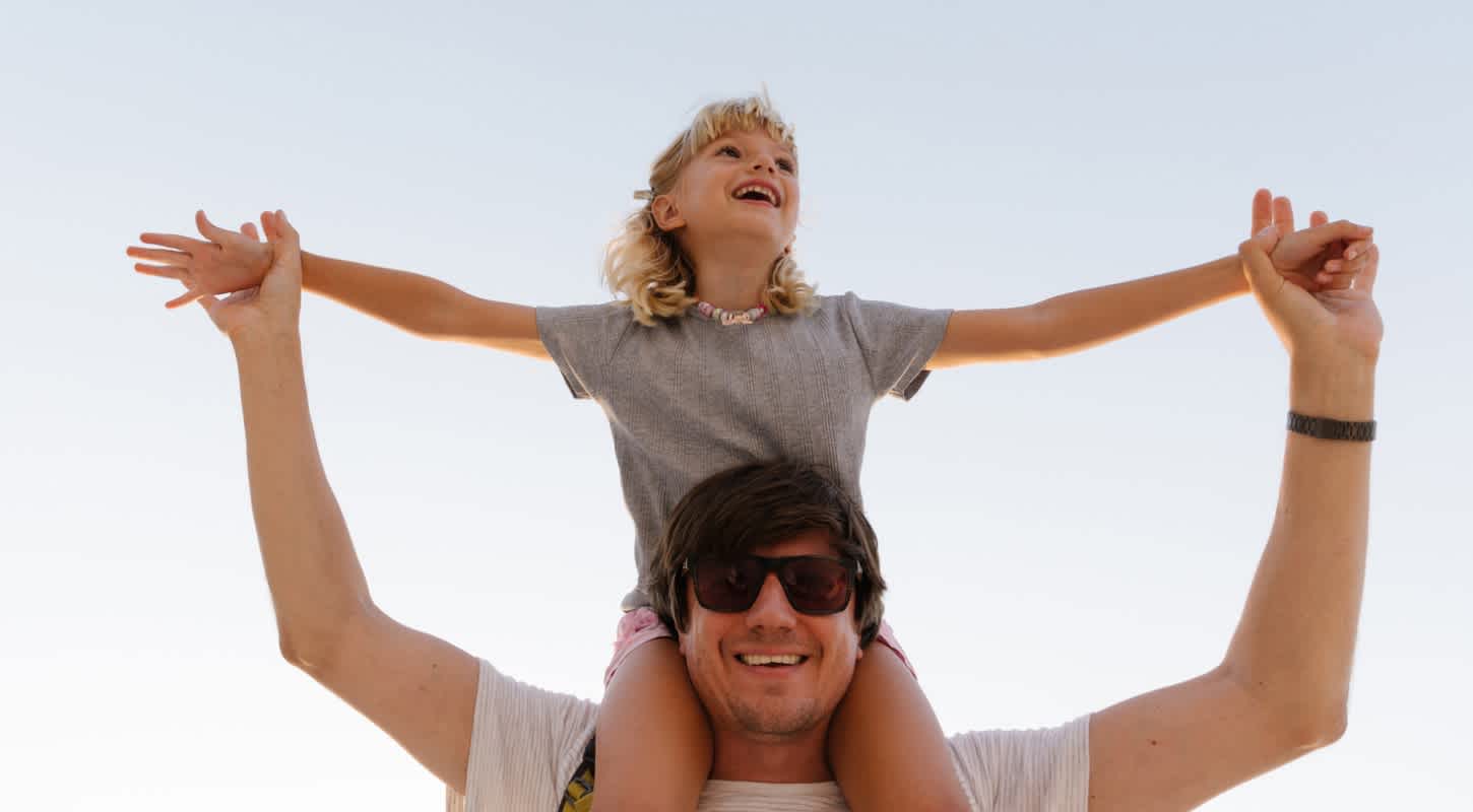 A toddler-aged girl sits atop her father's shoulders, her arms outstretched as he holds her hands. Both are laughing and smiling.