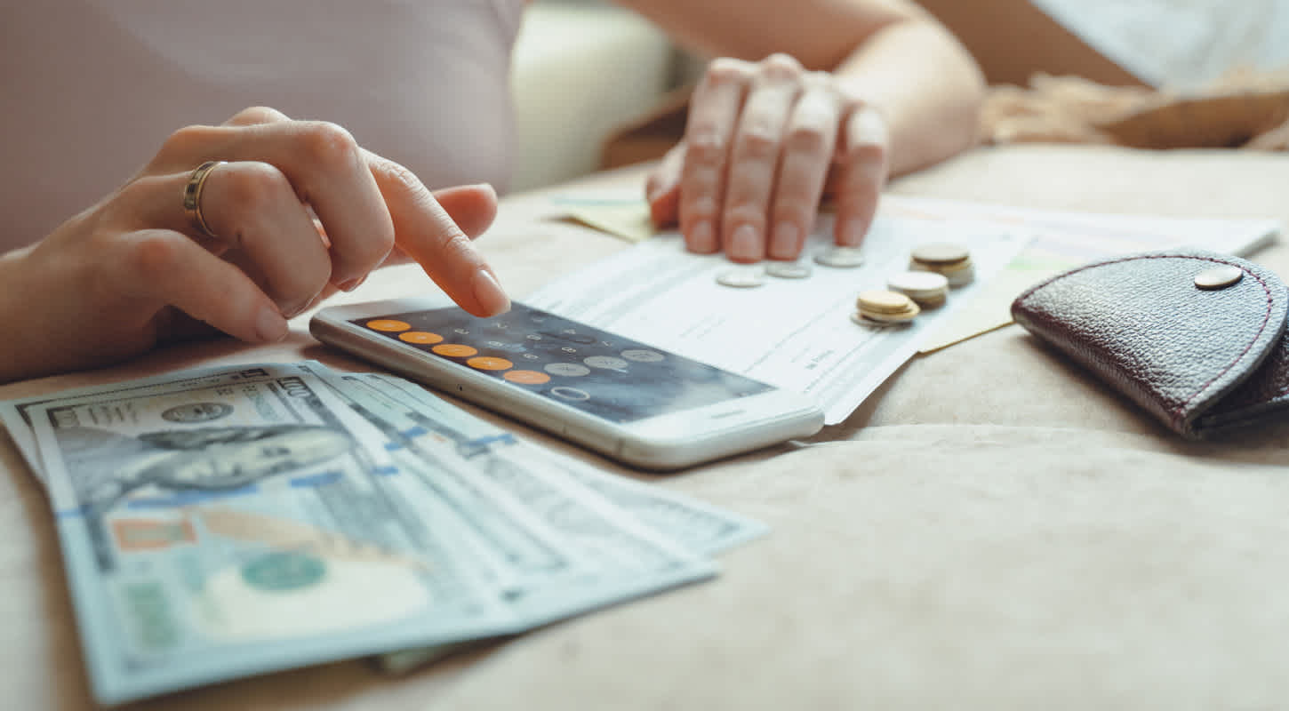 A woman uses her iPhone calculator to sum up the cash value of the dollar bills, coins, and checks on her desk space.
