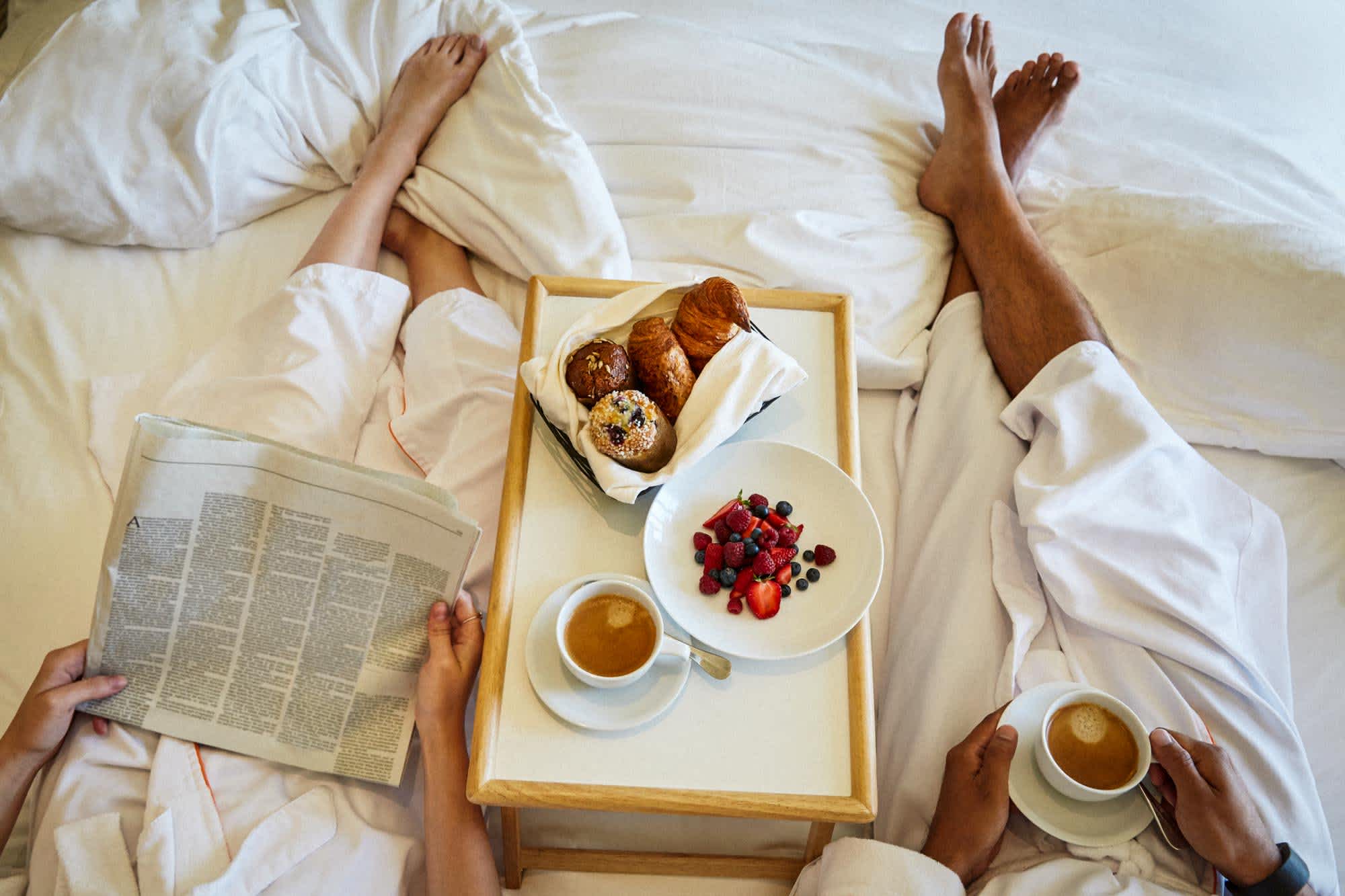 A couple lay in a plush white bed while enjoying morning room service. Mixed berries, croissants, muffins, coffee and a newspaper are at arms length.