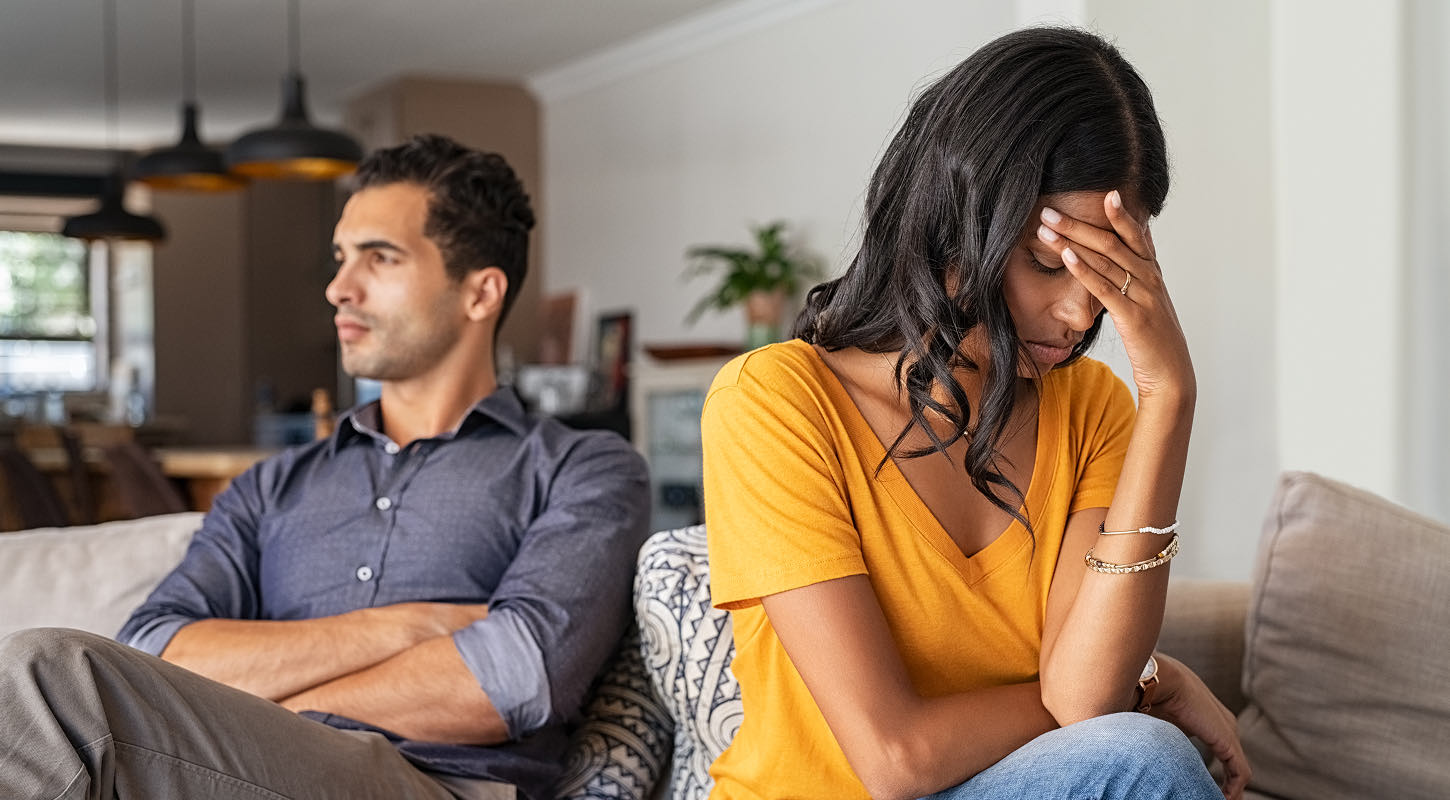 A young couple sits side by side on a couch. He sits with his arms crossed, staring into the distance. She rests her forehead on her hand, eyes closed in apparent dismay. 