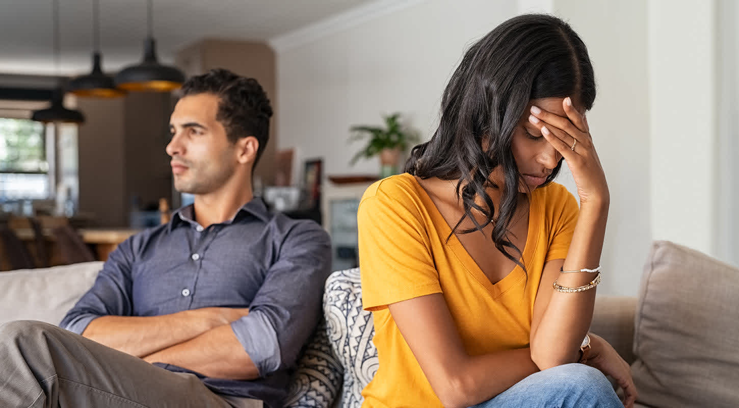 A young couple sits side by side on a couch. He sits with his arms crossed, staring into the distance. She rests her forehead on her hand, eyes closed in apparent dismay.