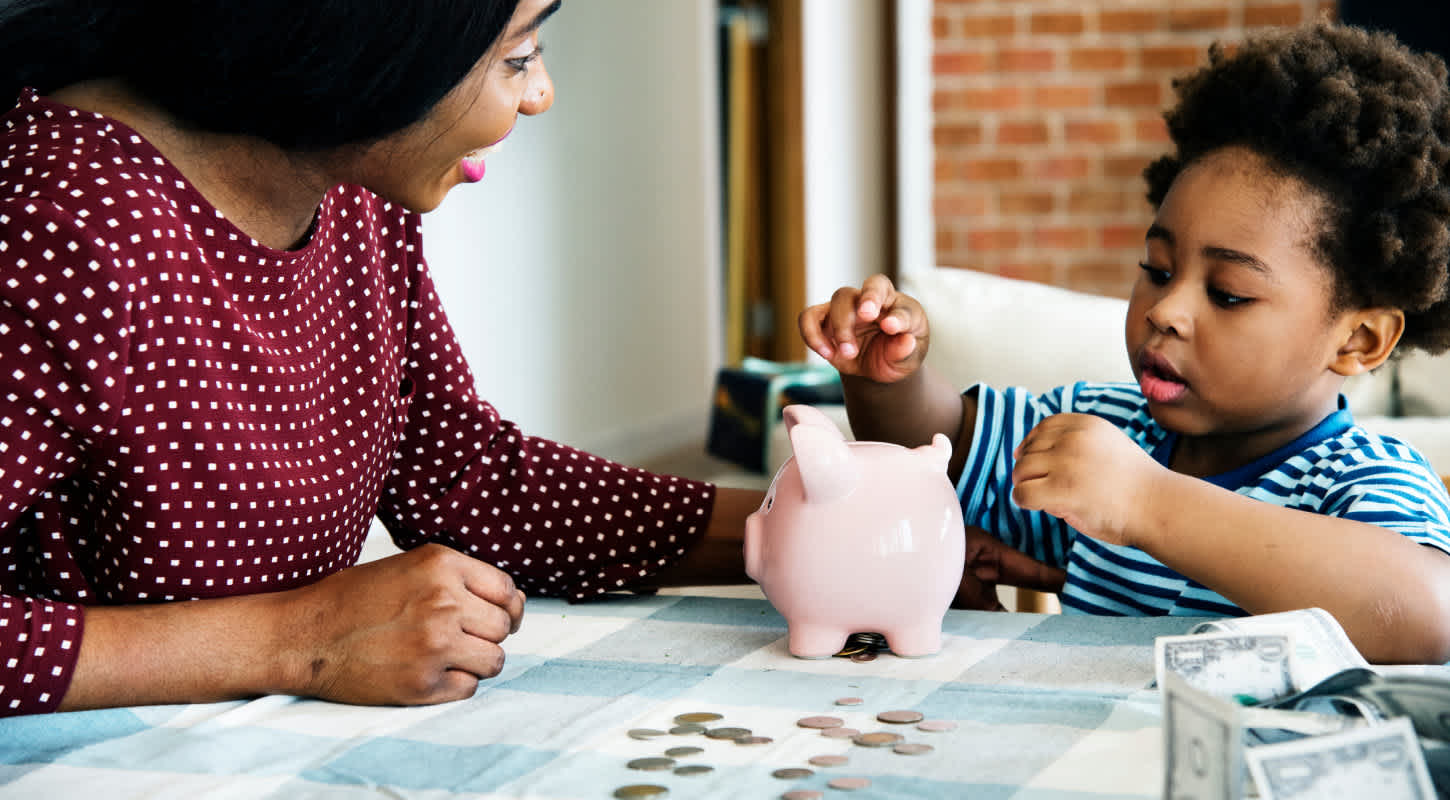 A mother watches over her toddler son as he saves coins inside of a piggy bank.