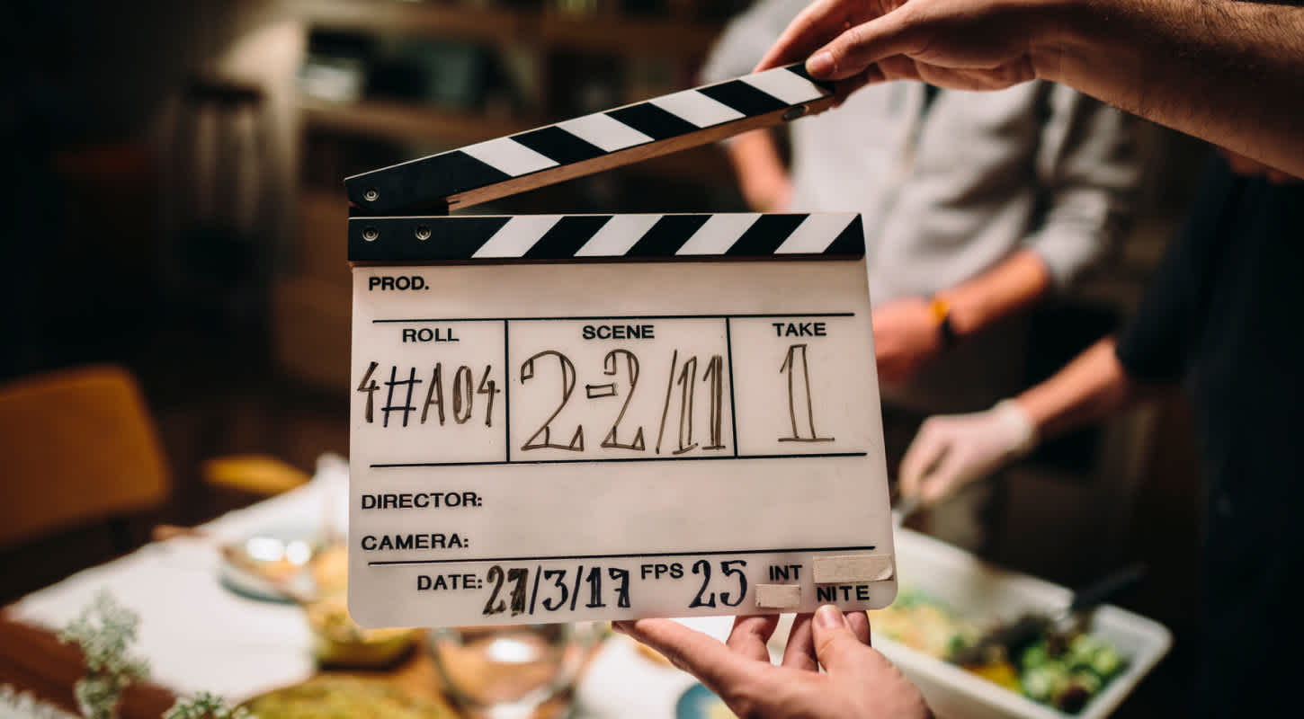 A clapperboard is held up by two hands in front of a table that has a spread of food served on it. Two individuals are serving themselves a meal from the buffet.