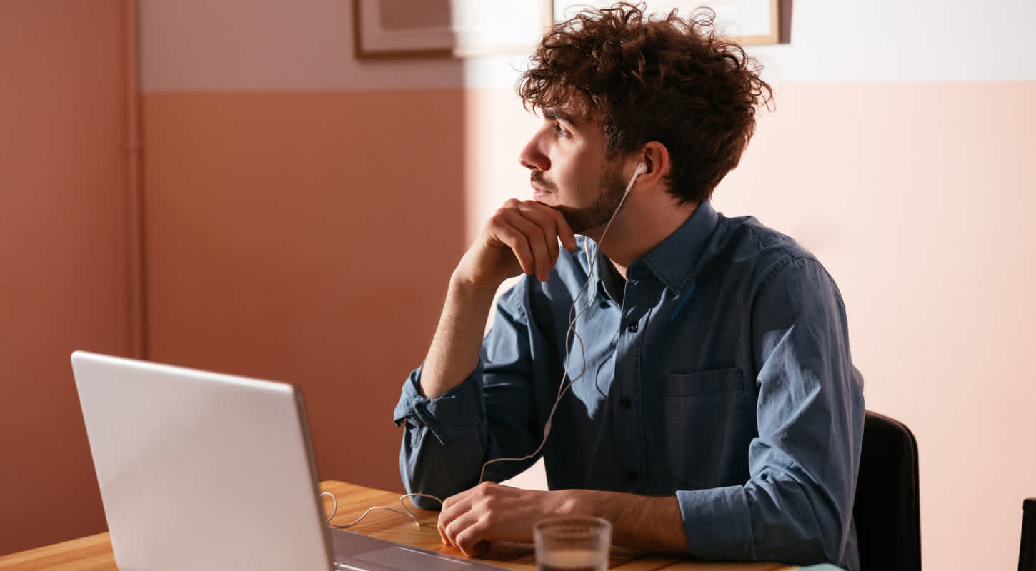A young man wearing headphones sits in front of his laptop with a cup of coffee, looking out the window as he ponders how much term life insurance he needs.