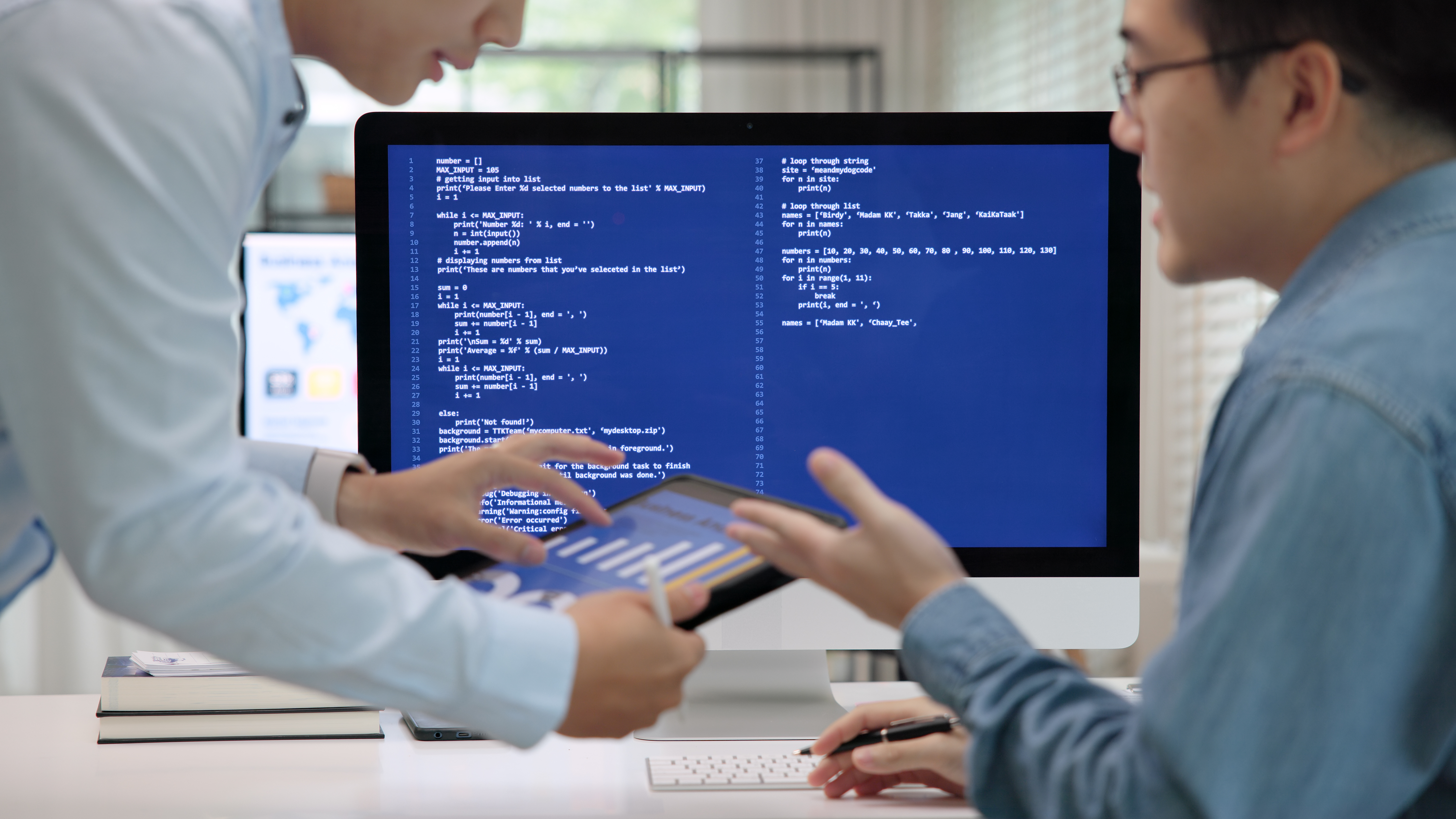 Two information technology employees are talking while looking at a tablet; it is implied that the man we see on the right of the frame is being trained by the other person, partially seen on the left. In the background is a computer screen depicting code.