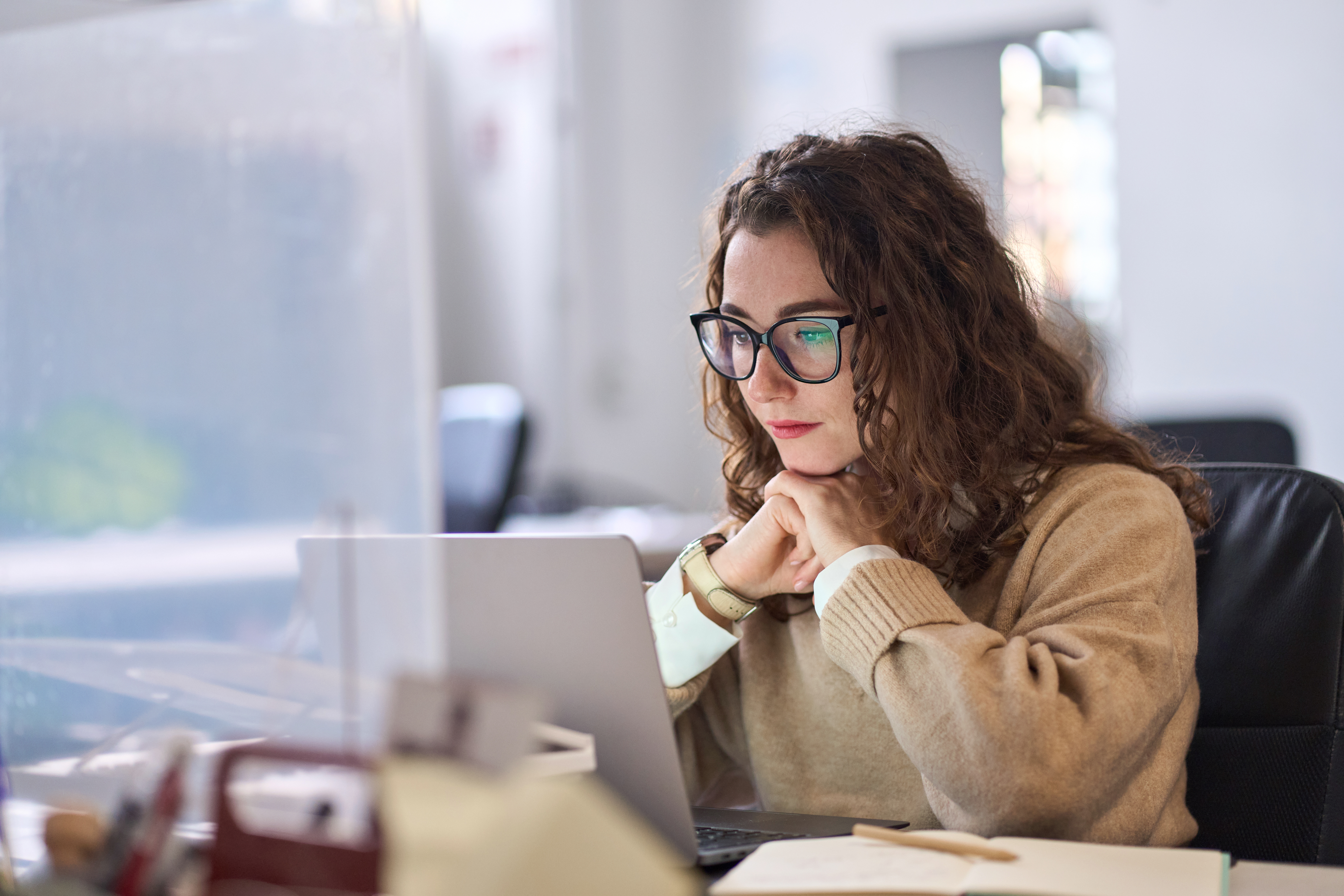 A woman wearing large glasses and a beige jumper sits at her desk completing training on her laptop. She is resting her chin on her hands.