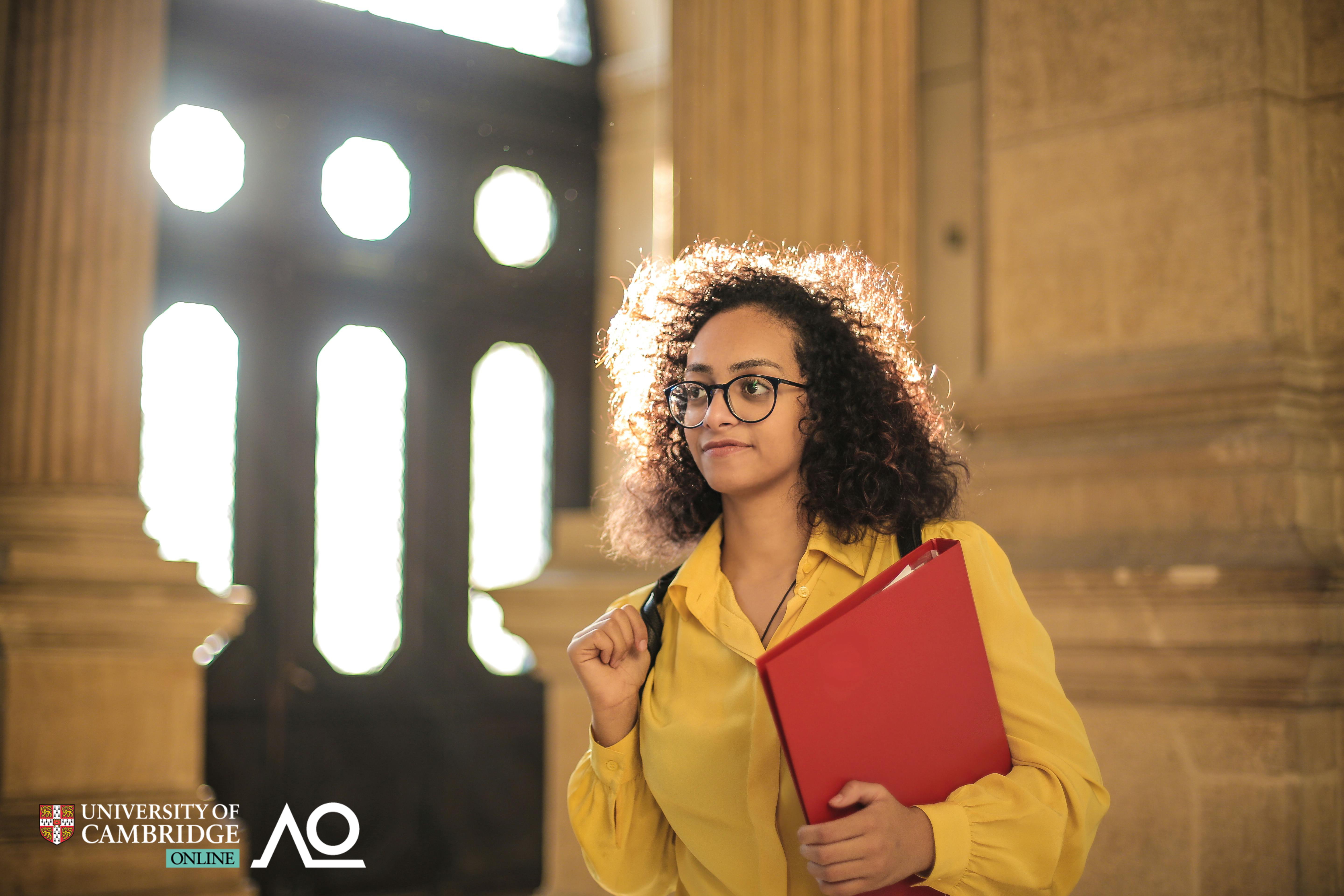 A young woman in a yellow coat is walking through the hall of an old building, carrying a red folder.