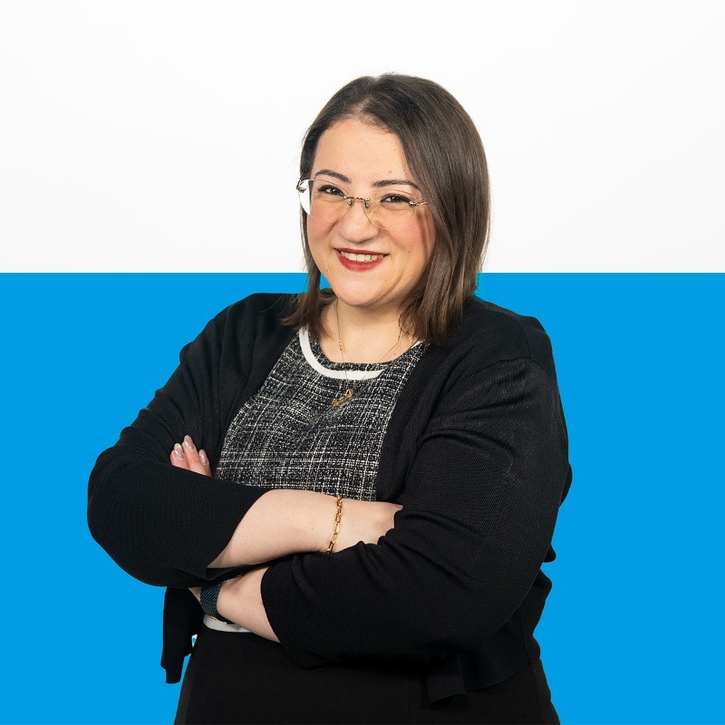 Portrait of Arwa Due-Gundersen against a blue and white background. She has short brown hair and transparent glasses, is wearing a black blazer and has her arms crossed in front of her.