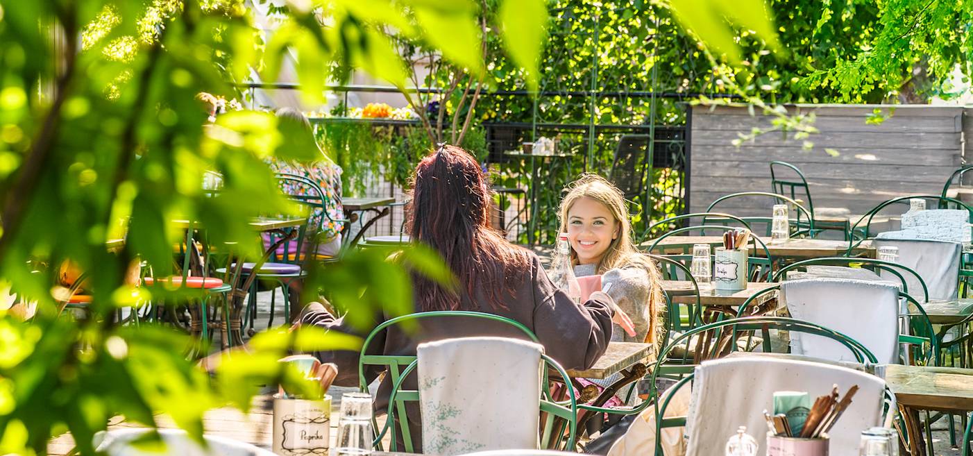 Two people sit at a table on a sunny outdoor café patio, surrounded by empty chairs and green plants. One person is smiling and holding a drink.