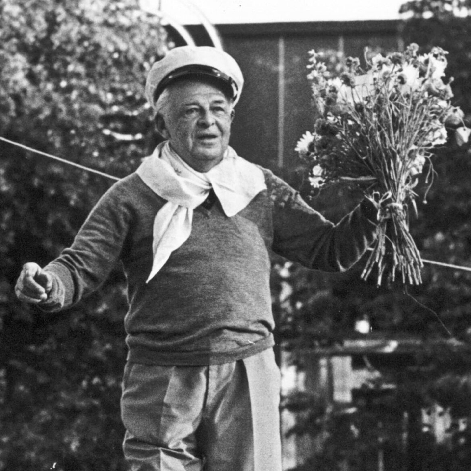 An older man in a helmet and scarf holds a bouquet of flowers while standing outdoors, with trees and a building in the background.