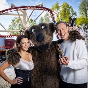 En kvinna och en man står bredvid en person i björndräkt på en nöjespark; mannen håller i en liten björnfigur och alla tre ler.