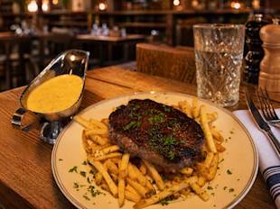 En grillad biff serverad på pommes frites, garnerad med örter, tillsammans med ett glas vatten och en portion gul sås på ett träbord i en restaurang.