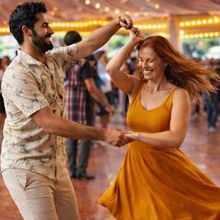 En man och en kvinna dansar tillsammans under ljusslingor på ett utomhusevenemang, ler och håller varandra i handen medan de snurrar.