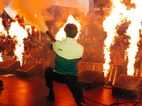 En artist på scenen står vänd mot en publik och håller i en dimmaskin med stora lågor som stiger upp runt scenen.