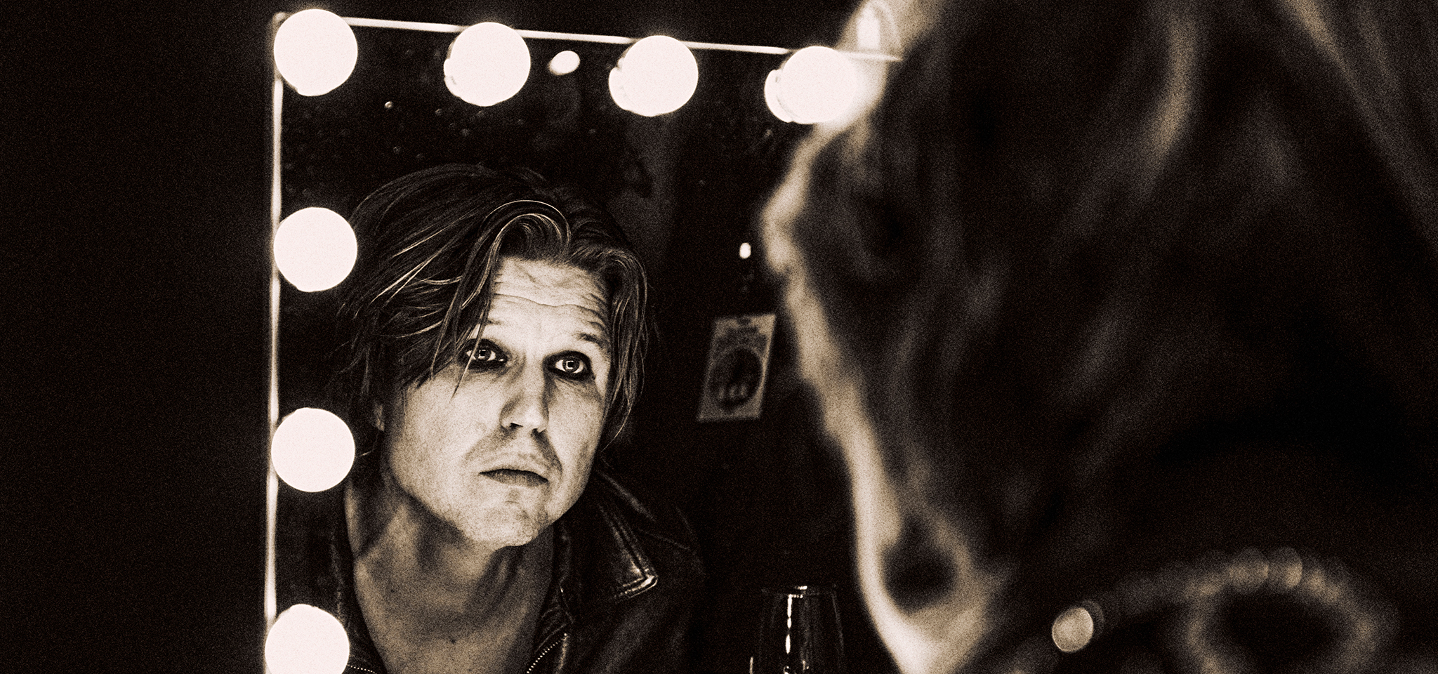 A person with shoulder-length hair and a leather jacket looks at their reflection in a mirror framed by lightbulbs in a dimly lit room.