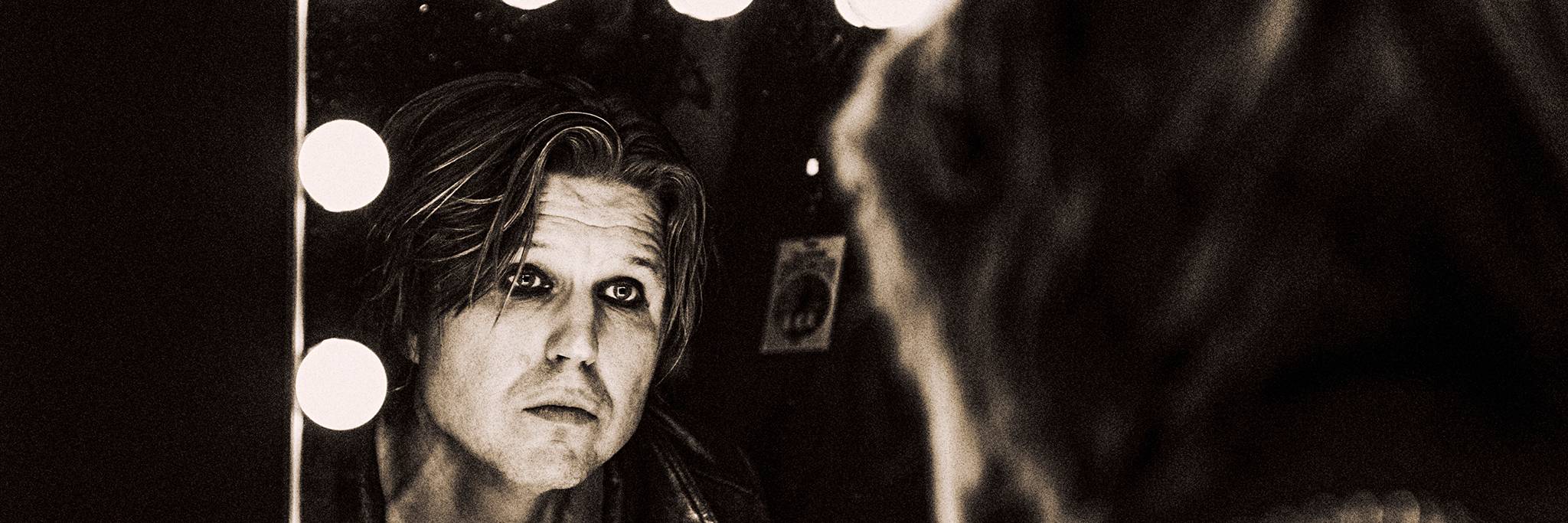 A person with shoulder-length hair and a leather jacket looks at their reflection in a mirror framed by lightbulbs in a dimly lit room.