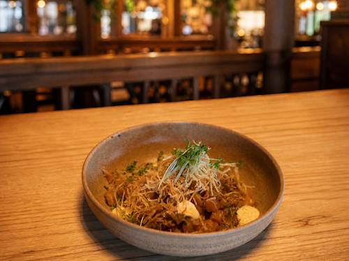 En keramikskål med en omsorgsfullt anrättad rätt, toppad med mikrogrönsaker och strimlad garnering, står på ett träbord i en varmt upplyst restaurang.