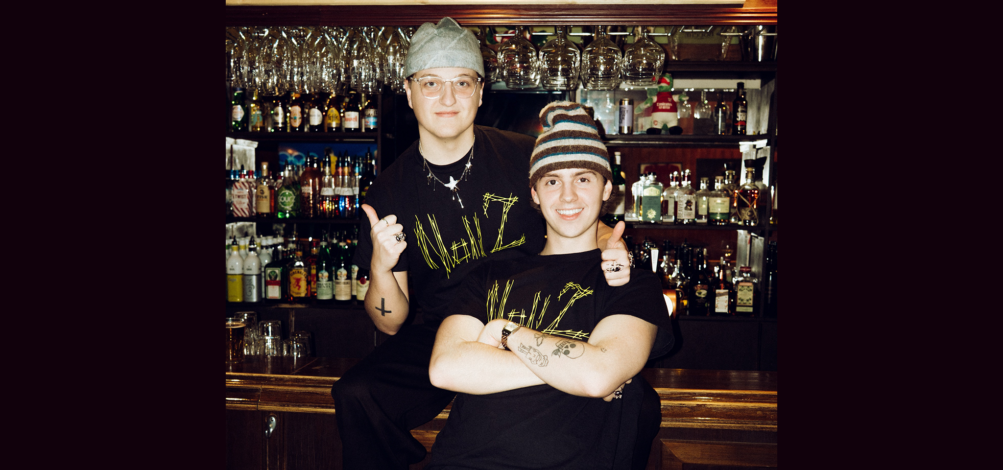Två personer i svarta t-shirts och hattar poserar tillsammans framför en bar fylld med flaskor och hängande glas. Den ena sitter med armarna i kors, den andra står bakom, ler och ger tummen upp.