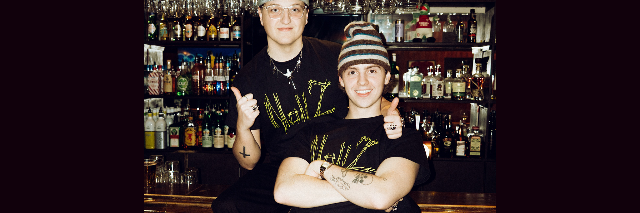 Två personer i svarta t-shirts och hattar poserar tillsammans framför en bar fylld med flaskor och hängande glas. Den ena sitter med armarna i kors, den andra står bakom, ler och ger tummen upp.