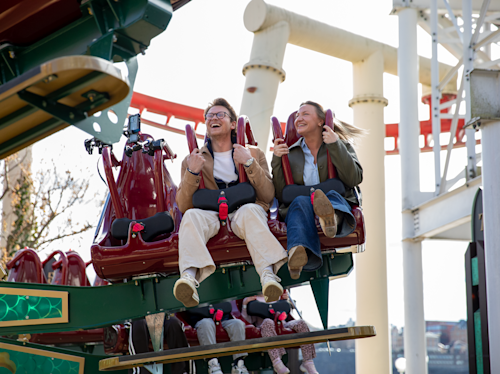 Två personer sitter sida vid sida på en åktur i ett nöjesfält, ler och håller i handtagen, med berg- och dalbanor och stålstöd synliga i bakgrunden.