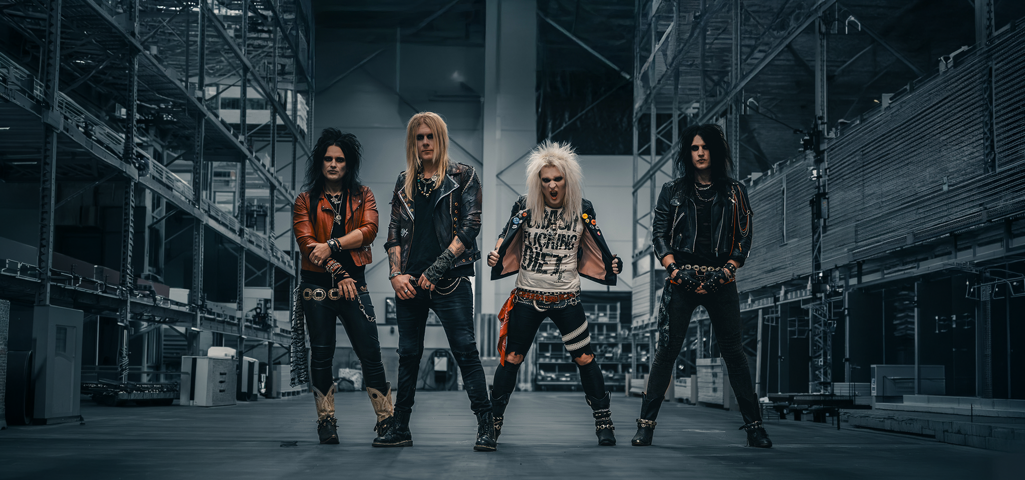 Fyra personer med långt hår och rockiga kläder står i en lagerlokal och tittar in i kameran med allvarliga ansiktsuttryck.