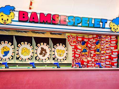 Colorful carnival booth with “BAMSESPELET” sign, spinning wheels featuring cartoon bear faces, and plush bear toys as prizes displayed on a red wall.