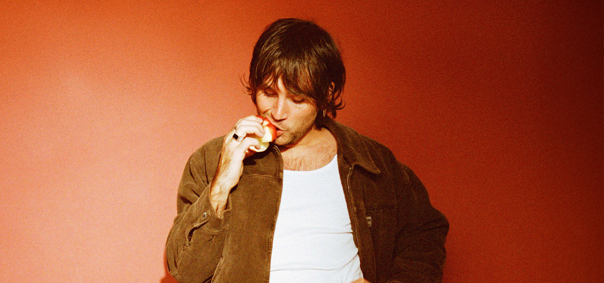 A person in a brown jacket and white shirt stands against a red background, eating an apple.