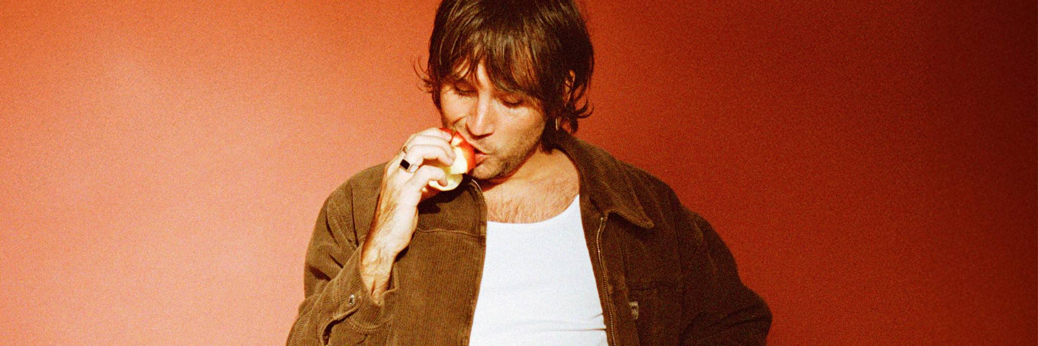 A person in a brown jacket and white shirt stands against a red background, eating an apple.