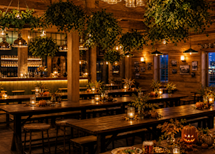 Rustic restaurant interior with wooden tables, hanging plants, warm lights, candles, autumn decorations, and plates of food and drinks on a table in the foreground.