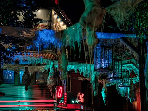 Street scene at night with buildings decorated in hanging cobweb-like material and eerie green and blue lighting, creating a spooky atmosphere.