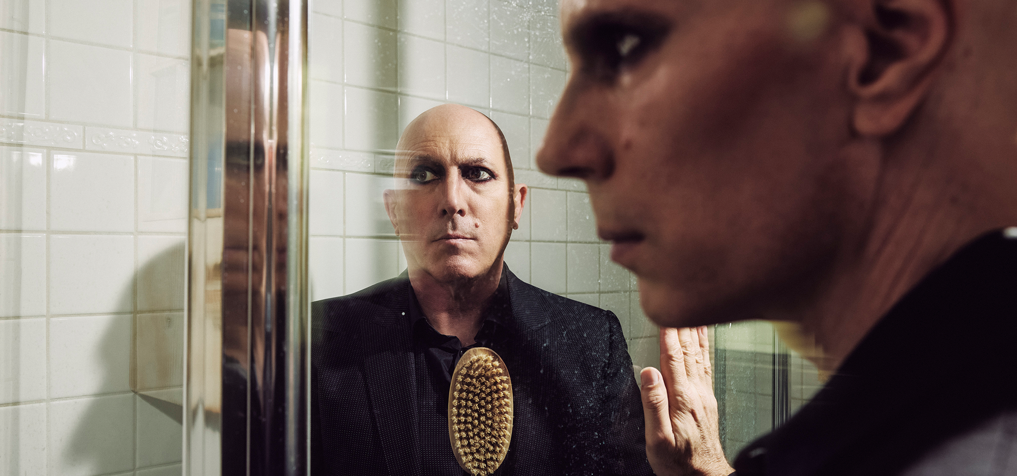 A bald person wearing dark clothing and eye makeup stands behind a glass shower door, looking at their own reflection.